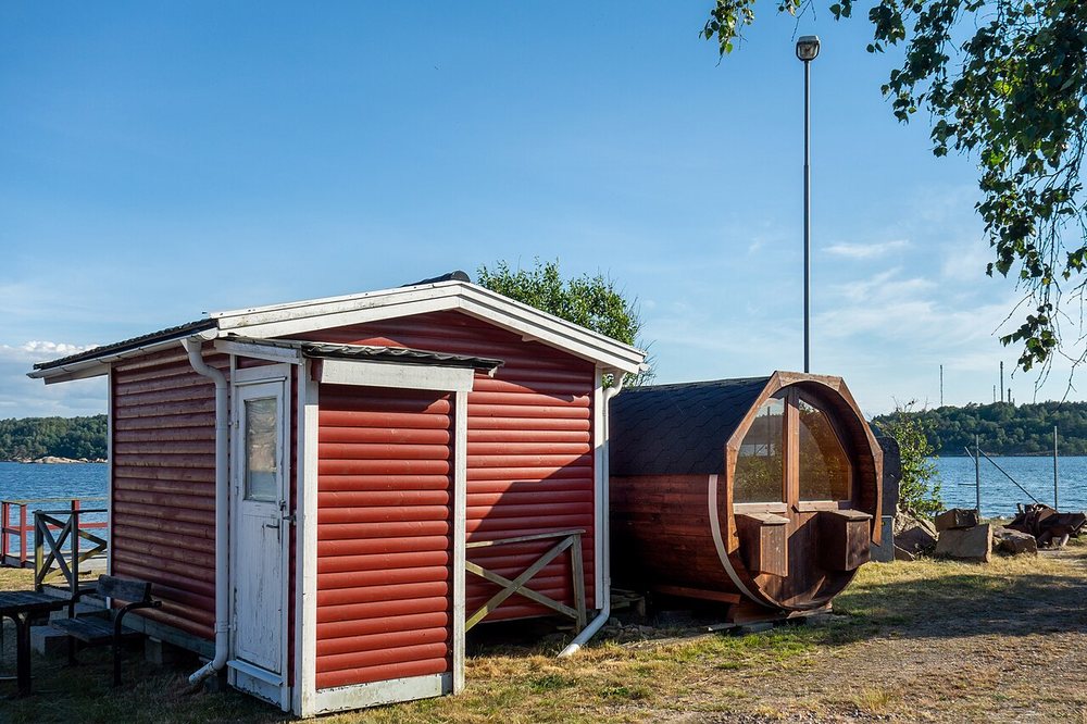 An outdoor wooden barrel sauna with a glass end wall next to a red cabin by the sea